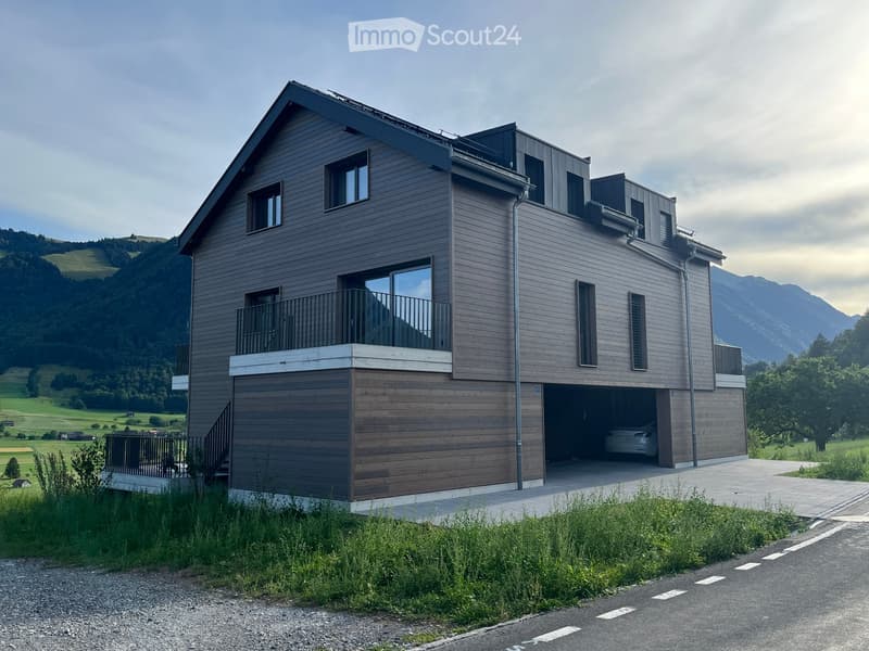 Modern two-story house with wooden siding, large windows, balcony, and garage, set in a scenic mountainous landscape.