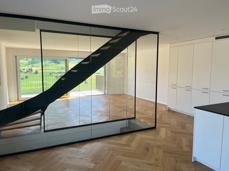 Modern hallway with wooden flooring, large mirrored wall, and a staircase leading upwards. The hallway features white cabinetry and a view of a green outdoor area through large windows.