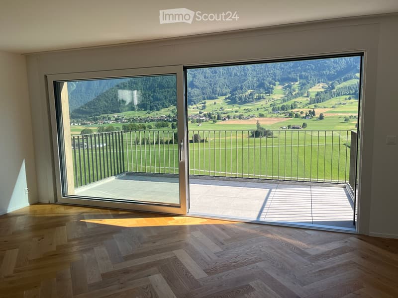 Open sliding glass doors lead to a spacious balcony with a metal railing, overlooking a scenic valley with green fields and distant mountains.