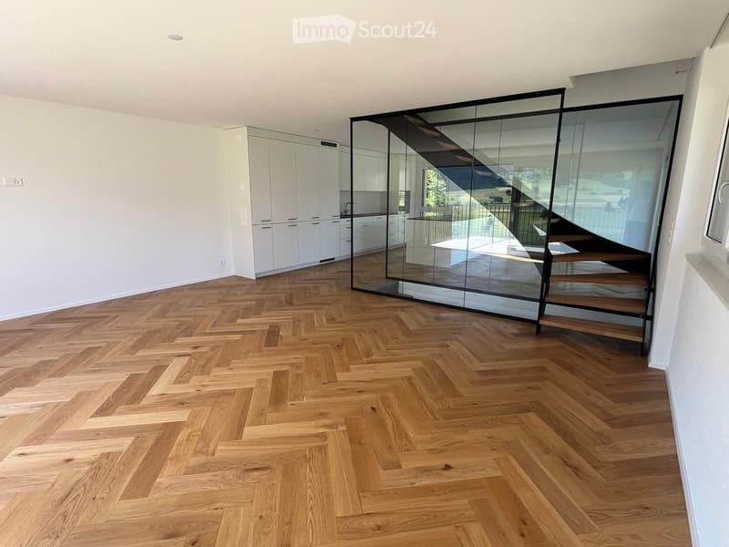 Open-plan space with herringbone wooden flooring, white walls, and a modern glass staircase leading to an upper level. The area includes built-in white cabinetry and a visible kitchen area in the background.