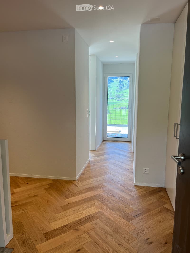 A modern hallway with wooden herringbone flooring, white walls, and recessed lighting. A glass door at the end leads to an outdoor area with a view of greenery.