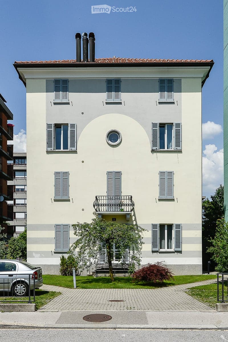 Three-story residential building with white facade, multiple windows with shutters, a small balcony on the second floor, and a tree in the front yard.