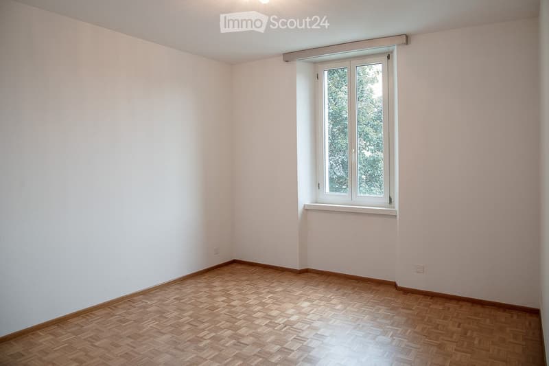 Empty room with light-colored walls and wooden parquet flooring, featuring a single window with white frame and blinds.