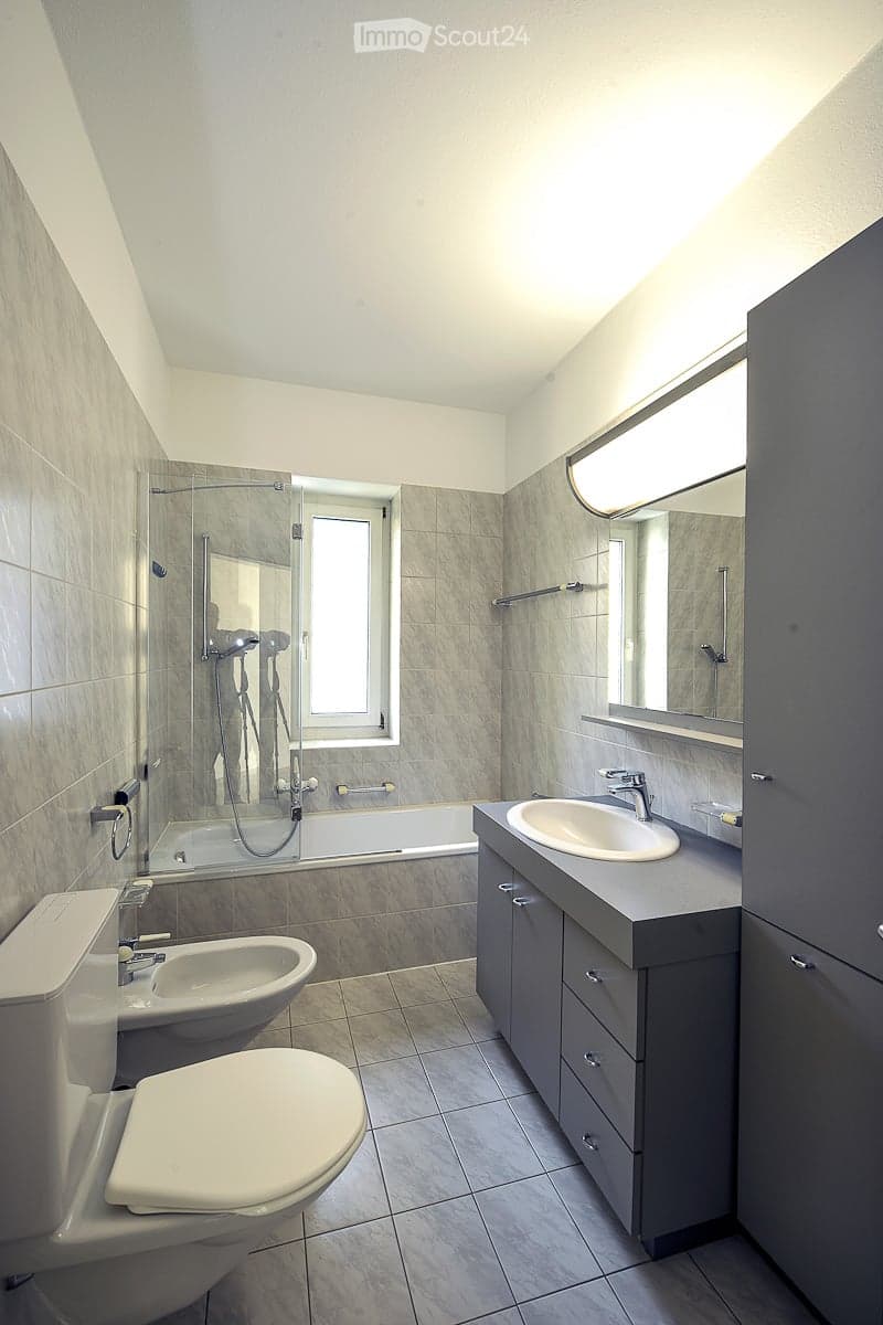 Modern bathroom with grey tiles, a bathtub, toilet, sink, and a large mirror above the sink. The shower area is enclosed with a glass door and has a window for natural light.