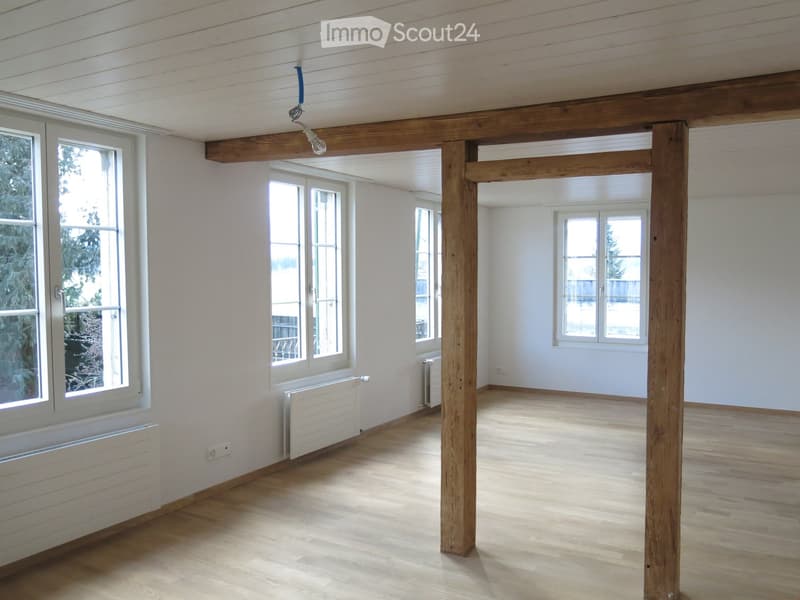 Empty room with wooden floor, white walls, large windows, and exposed wooden beams. Radiators are visible along the walls.