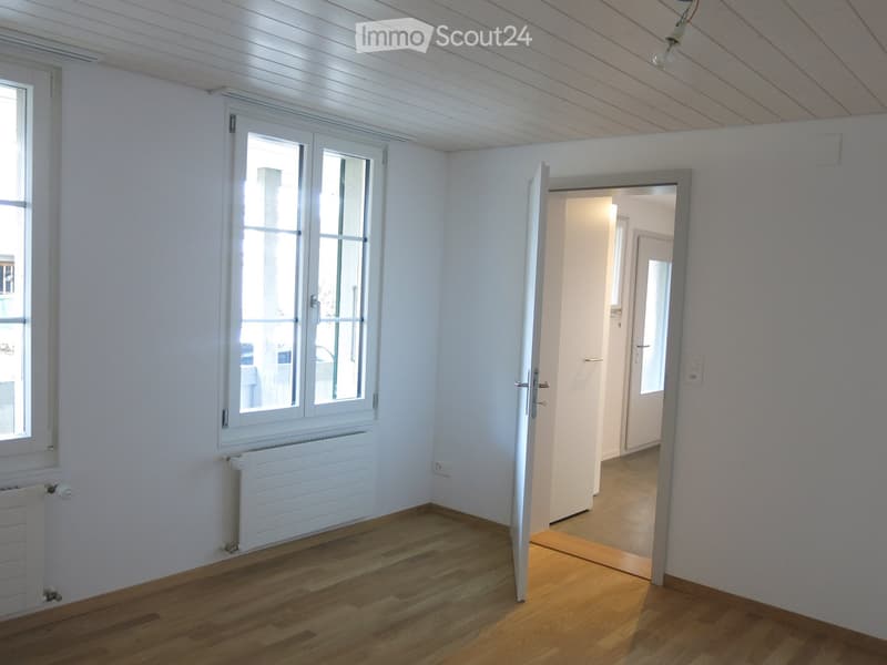 Empty room with light wooden flooring, white walls, and large windows. Radiators are visible under the windows, and an open doorway leads to another room.