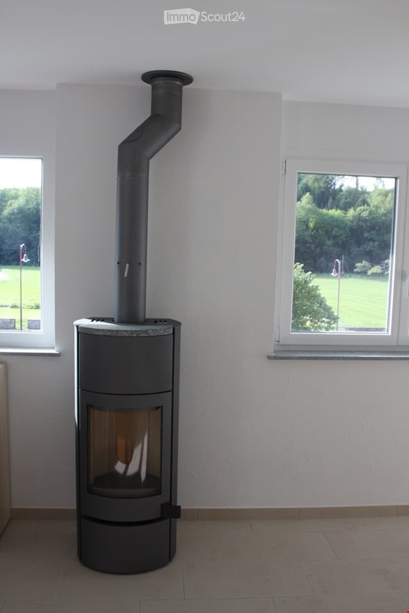 Modern wood-burning stove with a sleek black chimney, situated in a room with large windows and a tiled floor.