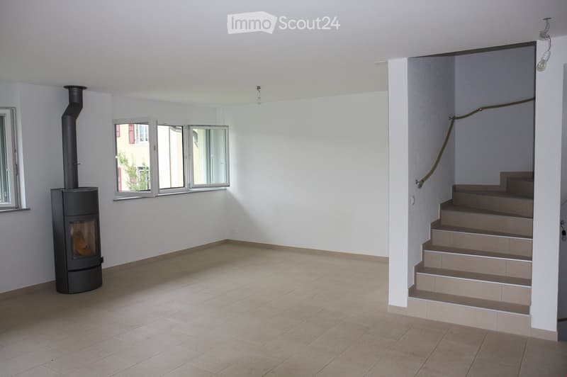 Empty room with tiled floor, white walls, a staircase leading upwards, and a wood-burning stove with a chimney.