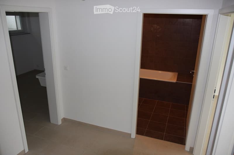 A narrow hallway with beige tiled flooring and white walls, leading to two rooms: a small bathroom with a toilet and a larger bathroom with a bathtub and tiled walls.