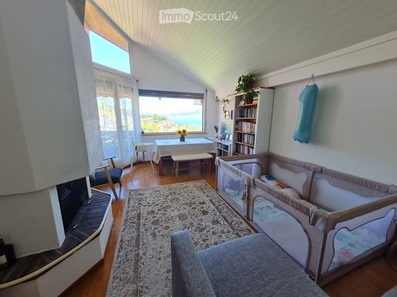 A cozy living room with a slanted ceiling, large windows offering a scenic view, a sofa, a coffee table, a bookshelf, a baby crib, and a rug.