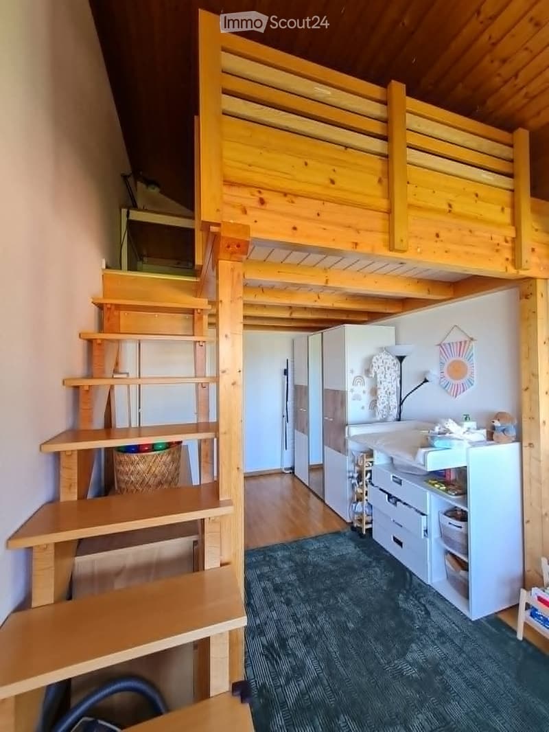 A compact, wooden-framed room with a loft bed, a small kitchen area, and a desk area. The kitchen includes a refrigerator, sink, and cabinets, while the loft bed is accessible via wooden stairs.