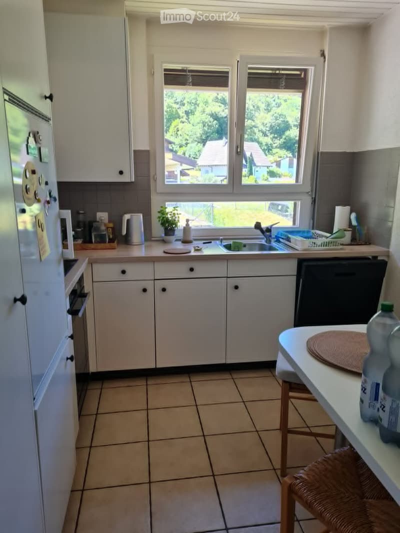 A well-lit kitchen with white cabinets, a tiled floor, and a window overlooking a green area. The kitchen features a sink, refrigerator, oven, dishwasher, and a small table with a chair.