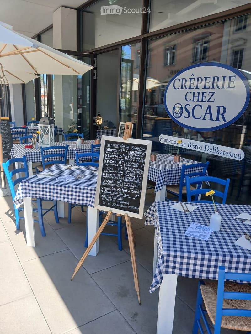Outdoor seating area of a restaurant with blue and white checkered tablecloths, blue chairs, and a menu board. The area is shaded by a large umbrella.