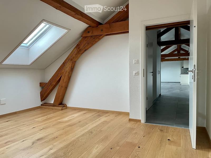 Empty attic room with wooden beams, skylight window, and wooden flooring. No furniture or equipment present.