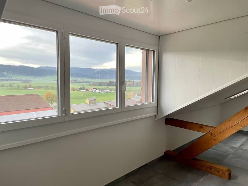 Bright hallway with large windows offering a scenic countryside view, wooden staircase leading upwards.