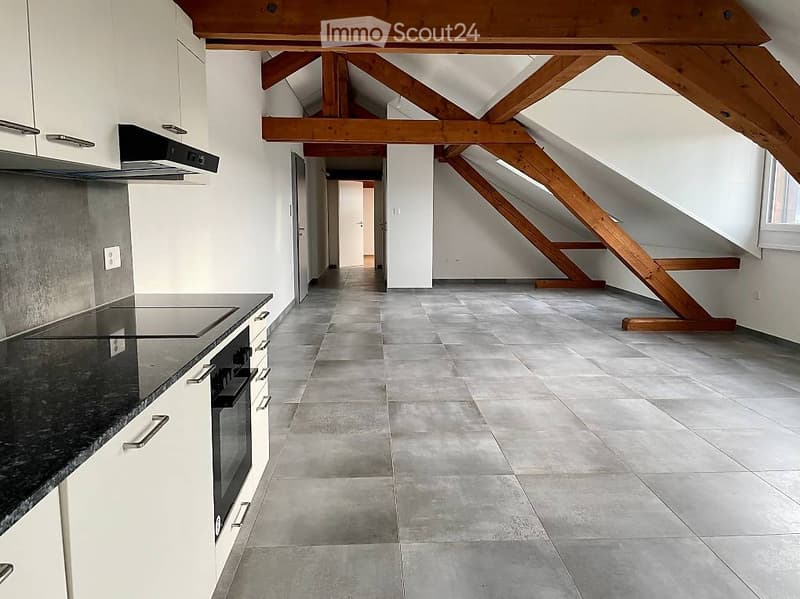 Empty attic space with exposed wooden beams, grey tiled floor, white walls, and a small kitchen area with cabinets and appliances on the left side.