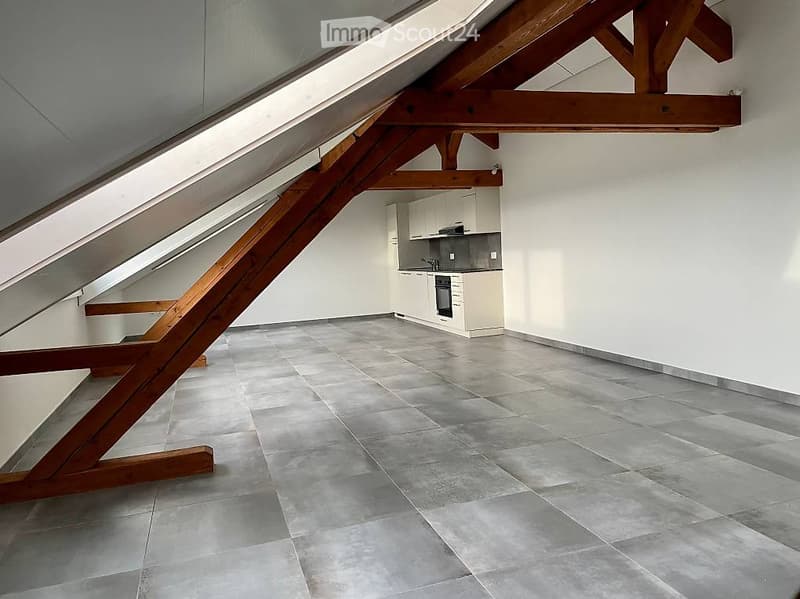 An attic space with exposed wooden beams, grey tiled flooring, and a small kitchen area featuring white cabinets and stainless steel appliances.