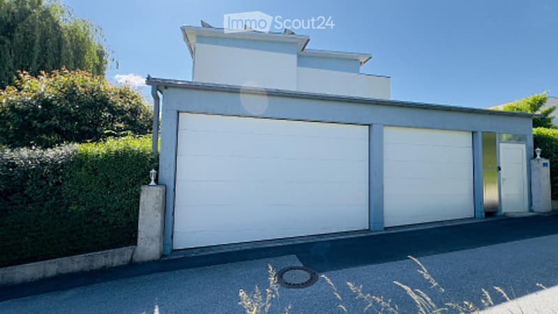 Modern two-story house with a large garage and a clean, contemporary design. The building features white walls, a flat roof, and is surrounded by greenery.