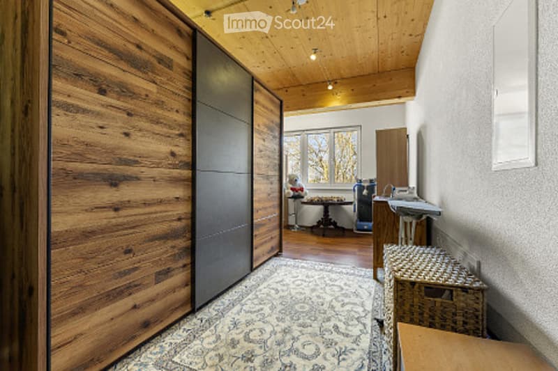 A hallway with wooden walls, a large wooden wardrobe, a patterned rug, a small table with a chair, and a stool near the wall.