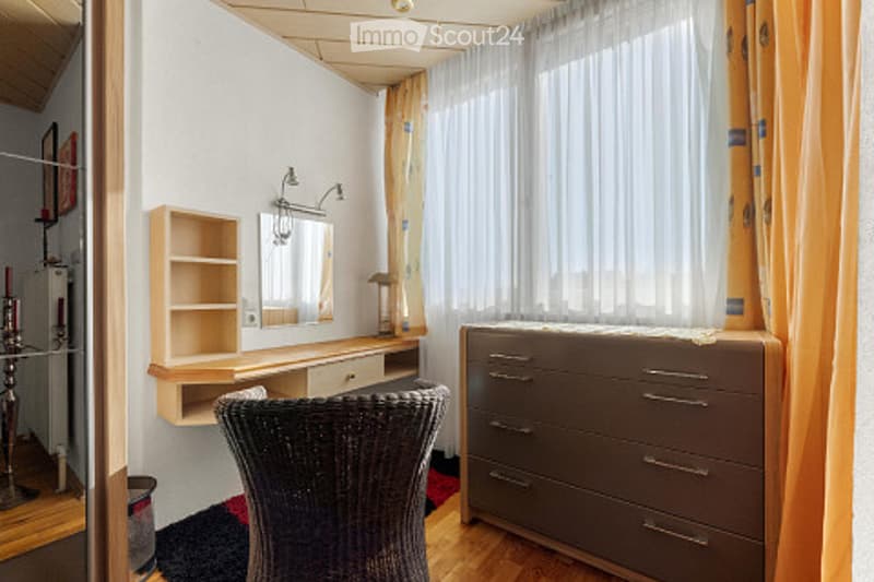 Cozy bedroom with wooden flooring, a large window with white curtains, a grey dresser, a wicker chair, a small shelf with a mirror, and a table lamp.