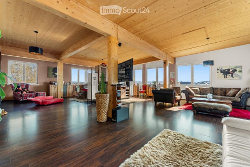 Spacious living room with exposed wooden beams, hardwood flooring, large windows, and a mix of seating including a sofa, armchair, and ottoman. A coffee table, floor lamp, and TV are present, along with decorative elements like a plant and picture frames.