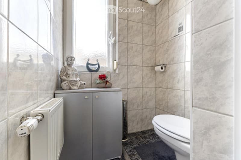 Modern bathroom with beige tiled walls, a toilet, a small cabinet with a radiator below, and a decorative statue on the cabinet.