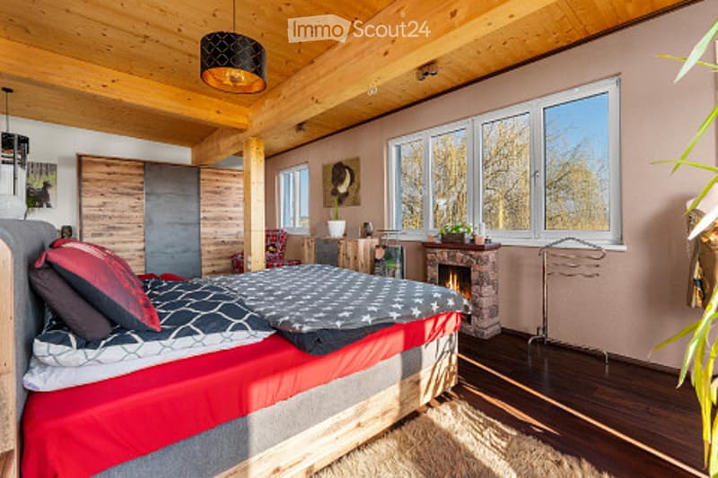 Cozy wooden bedroom with a large bed, pillows, blanket, nightstand, fireplace, and large windows letting in natural light.