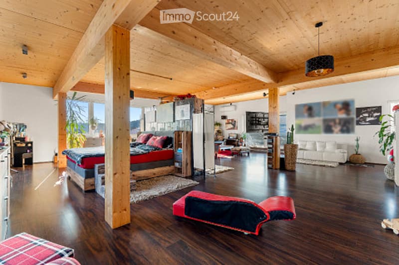 Open-plan living and bedroom area with wooden floors, exposed wooden beams, a large bed, a sofa, a table, and a lamp.