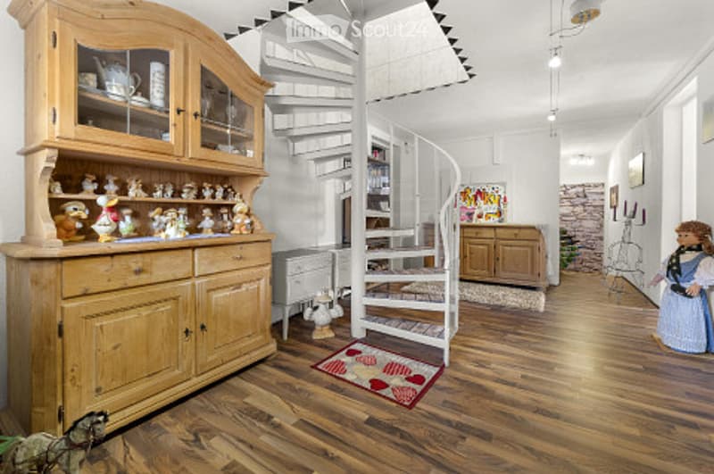 Bright, airy room with wooden flooring, a large wooden cabinet with glass doors, a spiral staircase, and a small table with chairs. A colorful rug with heart designs is placed near the staircase.