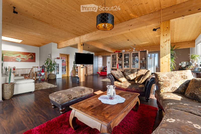 Spacious living room with wooden beams, hardwood flooring, large windows, a sofa, coffee table, ottoman, TV stand, and decorative plants.