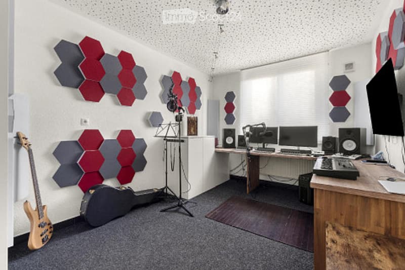 A modern, soundproofed music studio with acoustic panels on the walls, a guitar and case, a microphone stand, speakers, computer monitors, and a keyboard on a desk.