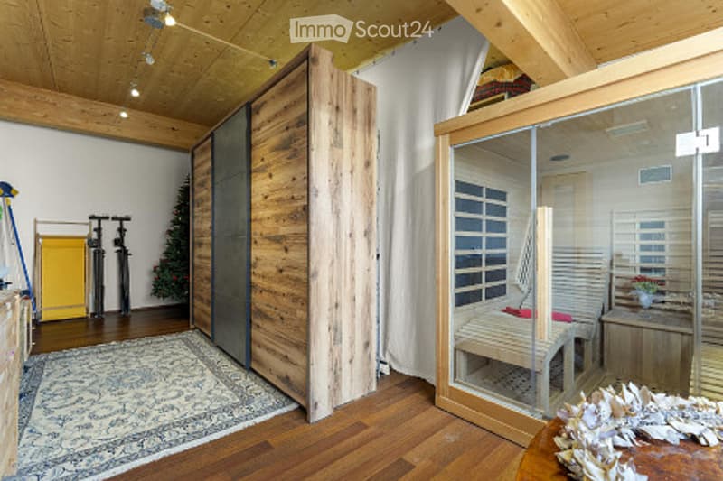 A modern, well-lit room with wooden floors and exposed beams. Features include a large wooden door, a glass-enclosed sauna with a bench and table, a patterned rug, and a small storage cabinet with a plant nearby.