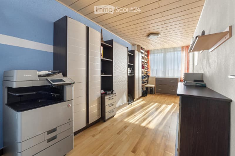 Bright, modern room with light blue walls, wooden flooring, and large windows. Features include a printer, built-in cabinets, shelves, a chair, a desk with a laptop, and additional storage units.