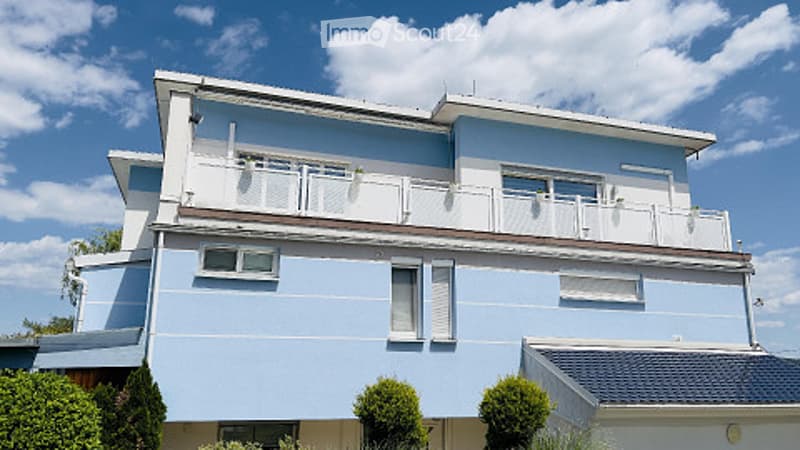 Two-story house with light blue exterior, white trim, multiple balconies, and neatly trimmed bushes in front.