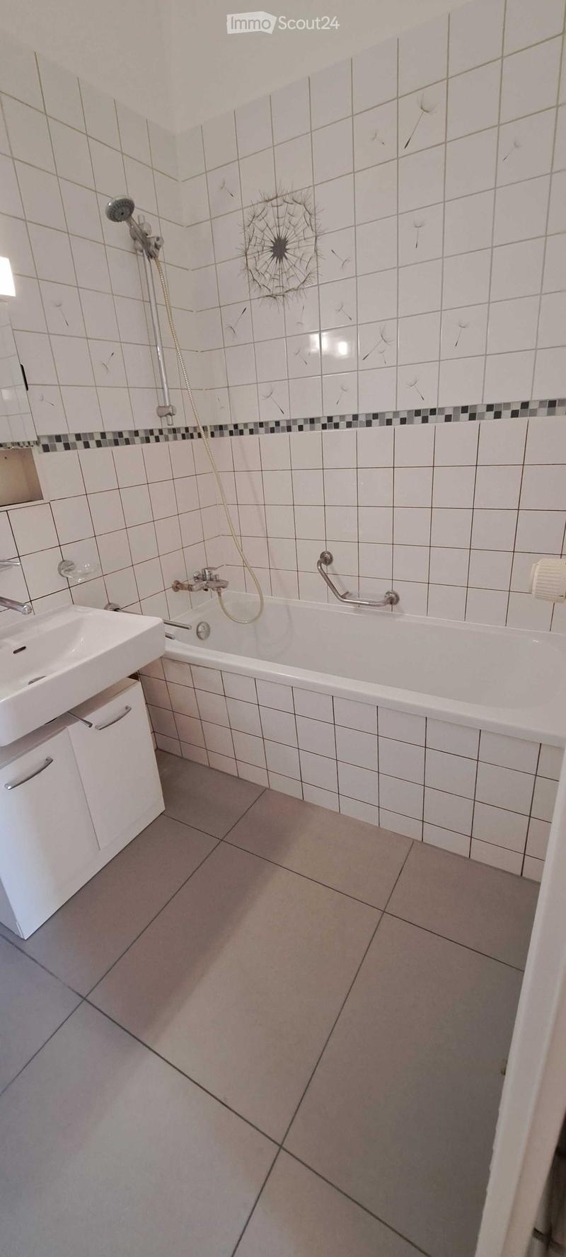 Bathroom with white tiled walls, a bathtub, a sink, and a showerhead. The tiles feature a dandelion design. The floor is tiled in a light gray color.