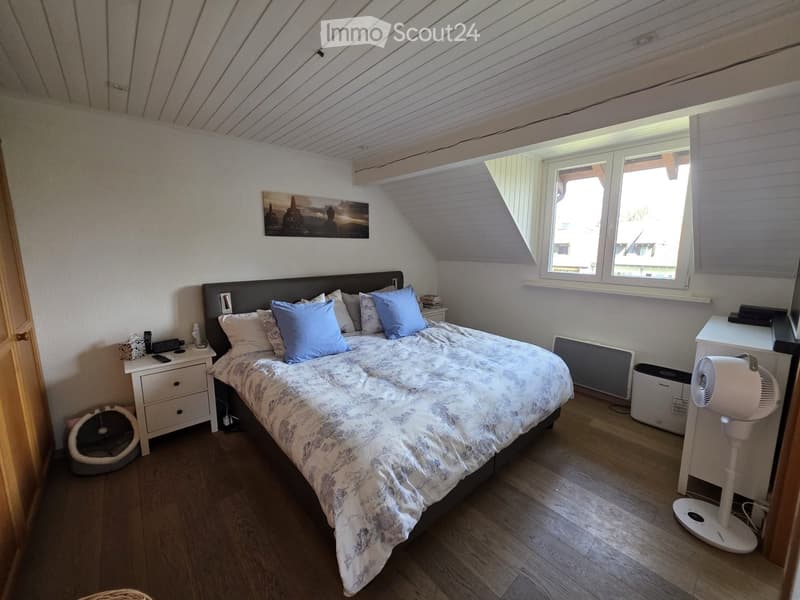 Cozy attic bedroom with wooden floors, slanted ceilings, large window, bed with pillows and blanket, nightstand, fan, and cat bed.