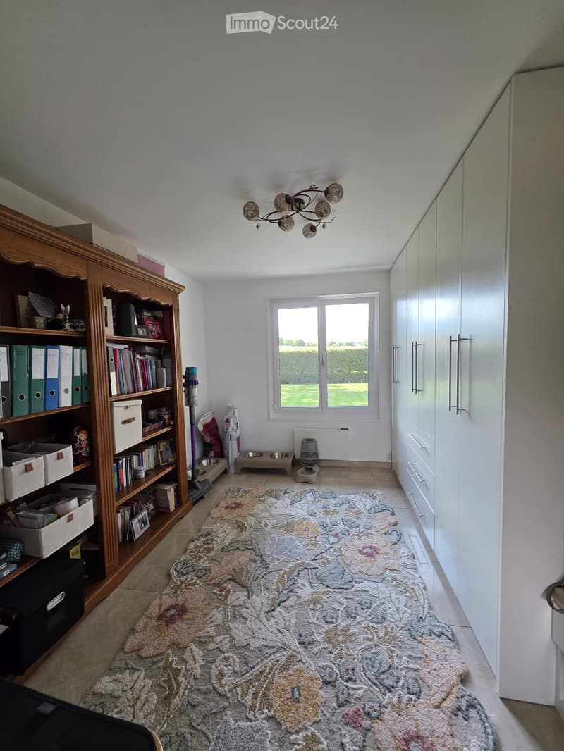 A well-lit hallway with a large patterned rug, wooden bookshelf filled with books and storage boxes, white cabinet, and cat supplies including food bowls and a litter box.