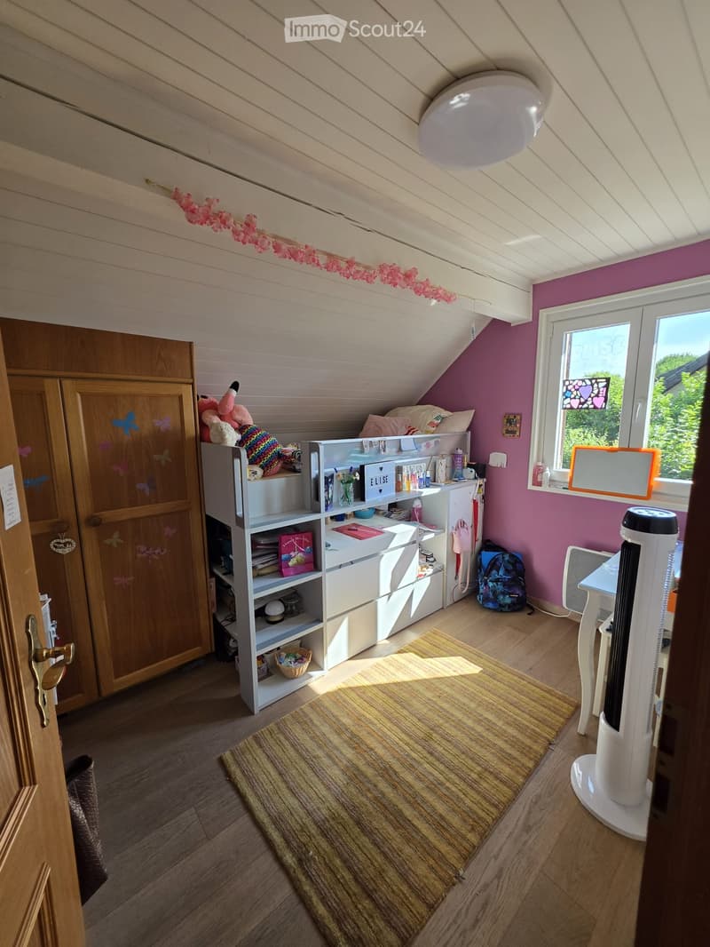 Cozy attic bedroom with sloped wooden ceiling, pink accent wall, bed with pillows, shelves filled with books and toys, fan, and a rug on the floor.