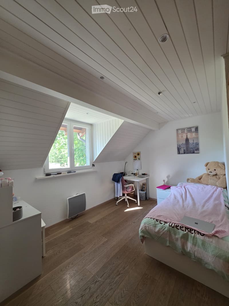 Cozy attic bedroom with wooden floors, slanted white ceilings, large windows, bed with pink and green bedding, desk with chair, teddy bear, and artwork on walls.