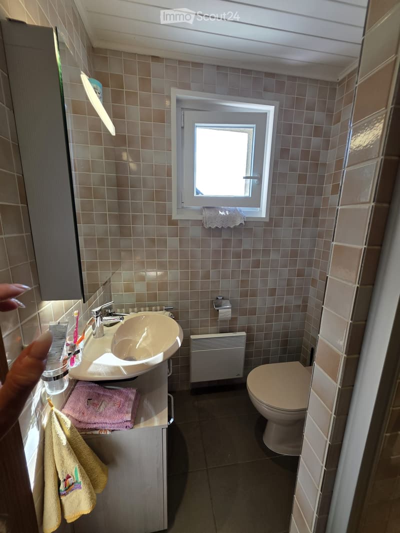 Small bathroom with beige tiles, a sink with a modern faucet, a toilet, a radiator, and a window with a lace curtain. Towels are placed on the sink cabinet.