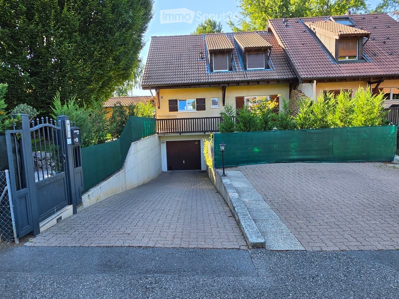 Two-story house with a red-tiled roof, multiple windows, and a garage. The property is surrounded by greenery and features a paved driveway leading to the entrance gate.