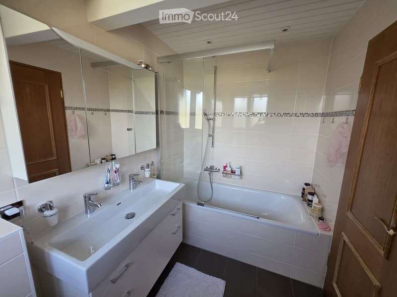 Modern bathroom with a large sink, bathtub, and shower area. Features include a large mirror, toothbrush holders, and various toiletries.