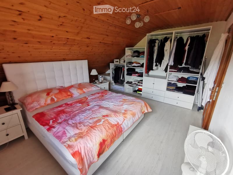 Cozy attic bedroom with wooden slanted ceilings, white bed with colorful bedding, nightstand with lamp, large wardrobe with clothes, and a fan in the corner.