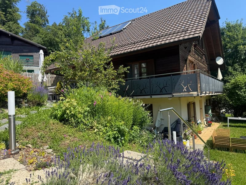 Two-story wooden house with a tiled roof, balcony, and lush garden with lavender plants and outdoor furniture.