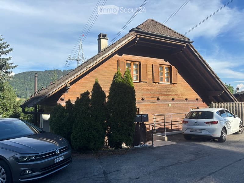 Two-story wooden house with a steep roof, multiple windows, and a small balcony. Two cars are parked in front of the house, and there are tall coniferous trees nearby.