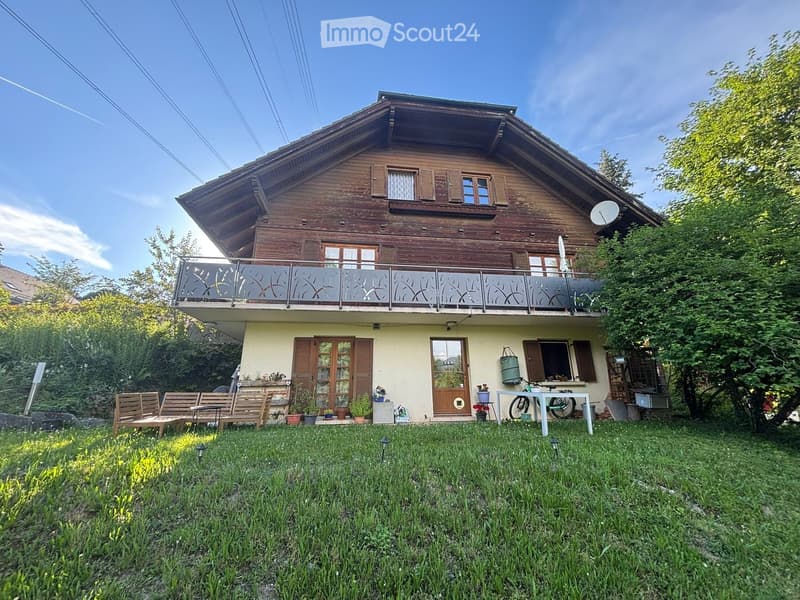 Two-story wooden house with a balcony, surrounded by a grassy lawn and garden area with outdoor furniture and plants.