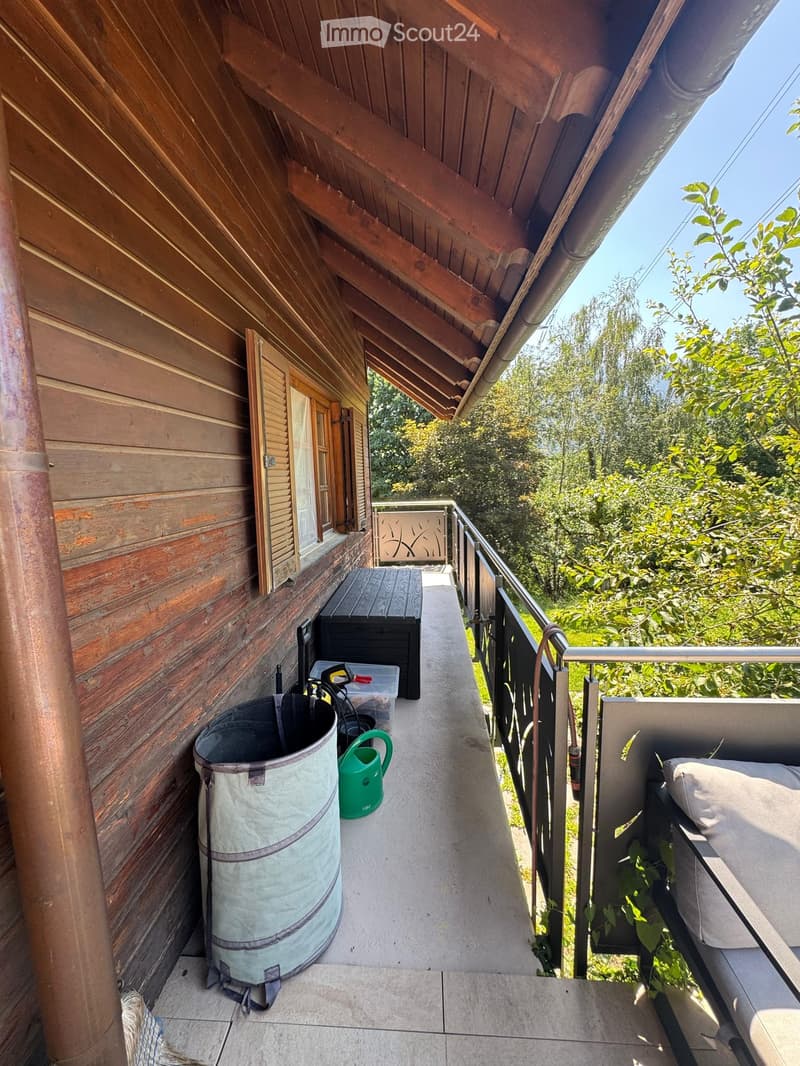 Balcony with wooden walls, metal railing, and a view of greenery. Includes a stool, trash can, cup, and part of a sofa.
