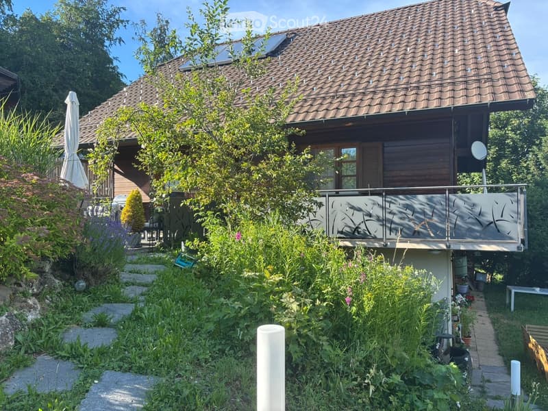 A two-story house with a brown tiled roof, surrounded by lush greenery and a well-maintained garden. The house features large windows, a balcony with a modern railing, and a patio area with outdoor furniture.