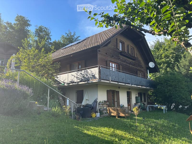 A two-story wooden house with a balcony, surrounded by greenery and a well-maintained lawn.