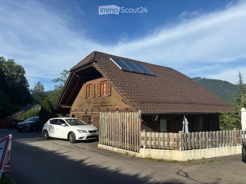 A two-story wooden house with a brown tiled roof, solar panels, and a wooden fence in front. Two cars are parked in front of the house.